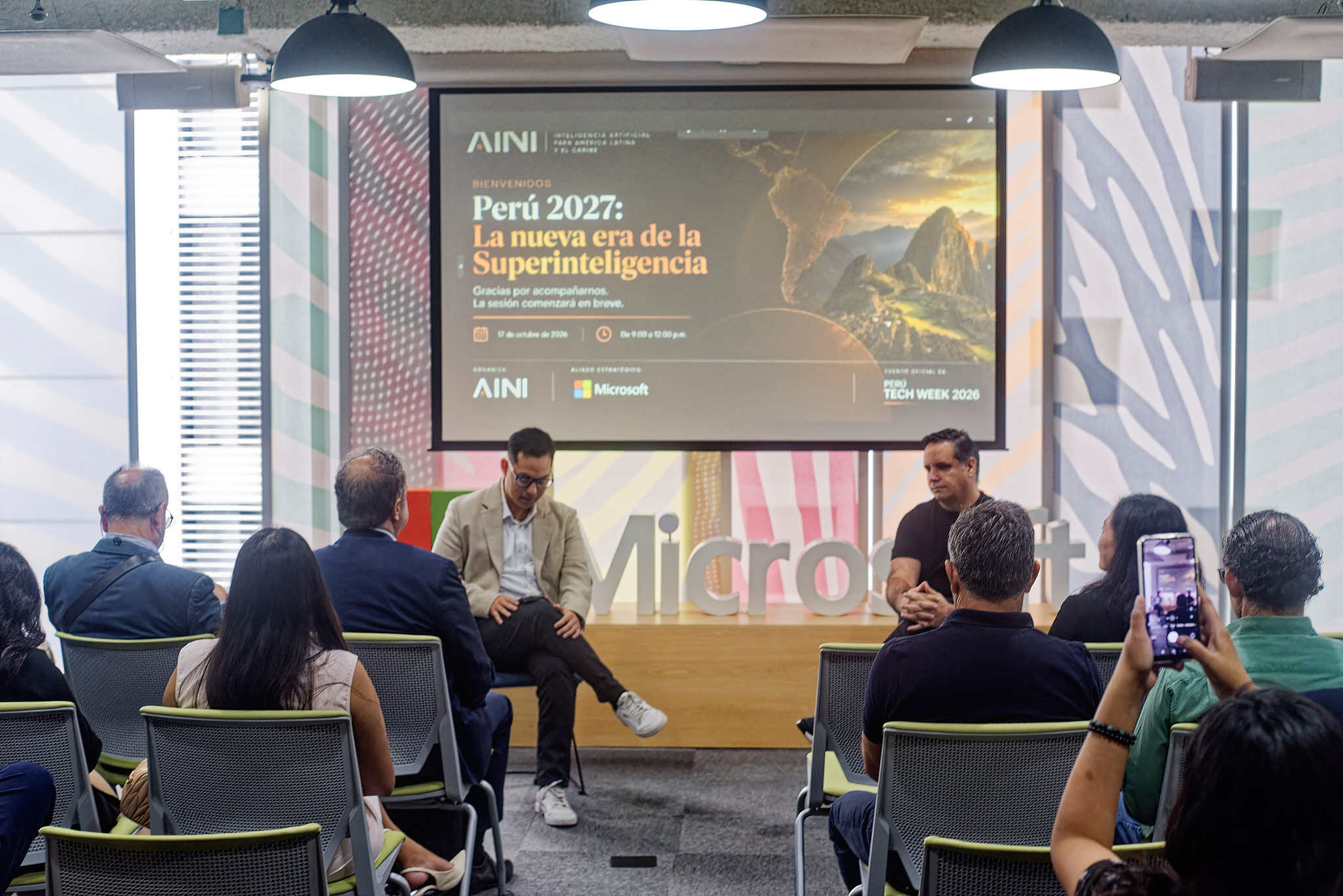 Two speakers talk seated on stage, in front of the event screen and the Microsoft sign, with the audience seen from behind in the foreground